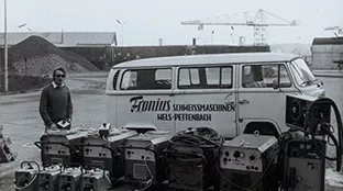 A white bus with the words “Fronius Schweißmaschinen Wels-Pettenbach” (Fronius Welding Machines Wels-Pettenbach) written on it, with a row of welding machines in front of it.