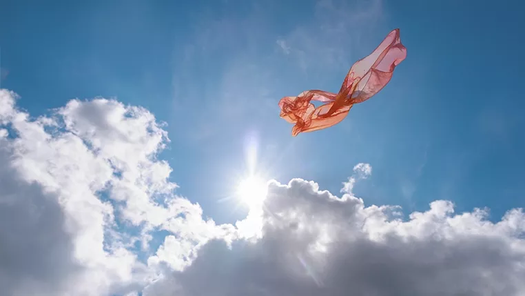 Ein Stück rosafarbenes, transparentes Stofftuch schwebt vor blauem Himmel mit Sonne und weißen Wolken.