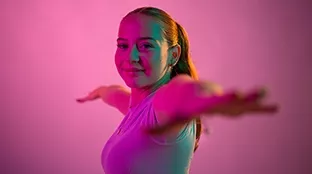 Junge Frau streckt ihre Arme aus und lächelt vor einem pinken Hintergrund in die Kamera.
