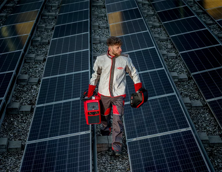 In the center is a man in uniform with a Fronius welding machine, walking along a roof covered with solar panels.