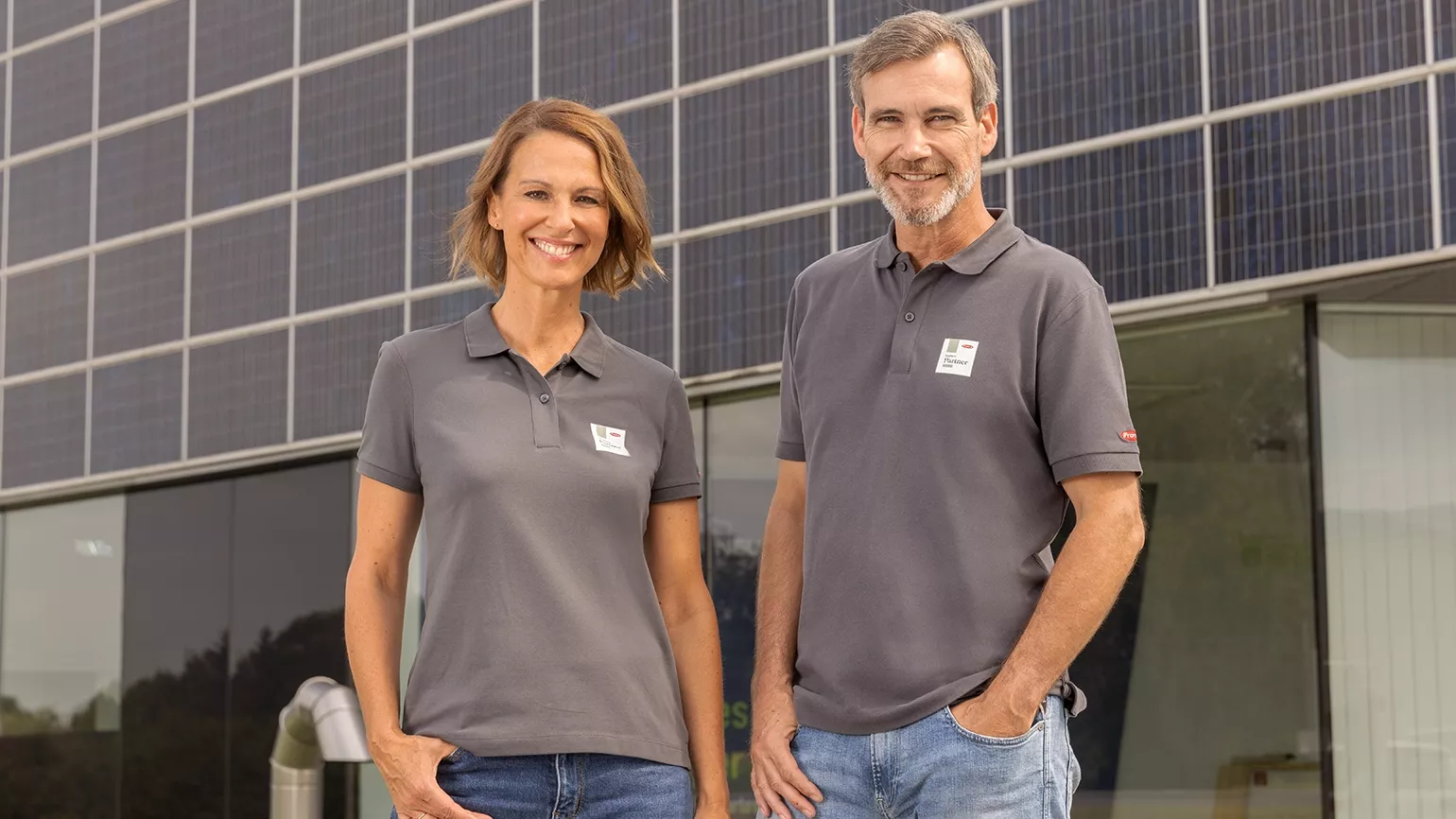 Two people wearing grey polo shirts standing in front of a large photovoltaic system on a modern building.