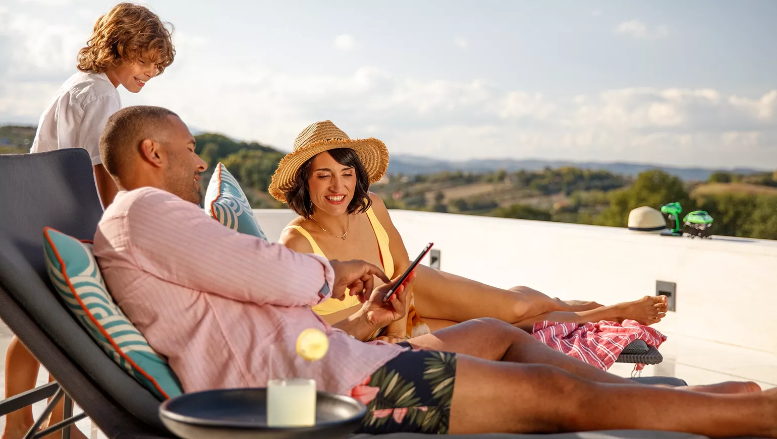 Two adults and a child relaxing on a terrace with scenic hills in the background; one person holding a tablet, a small table with a drink nearby.