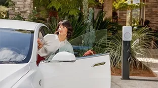 Eine Frau steigt in ihr Elektroauto ein, im Hintergrund ist eine Fronius Wattpilot Flex Wallbox zu sehen.