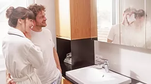 Two people standing together in a bathroom in front of a sink, with a heart drawn on the fogged-up mirror.